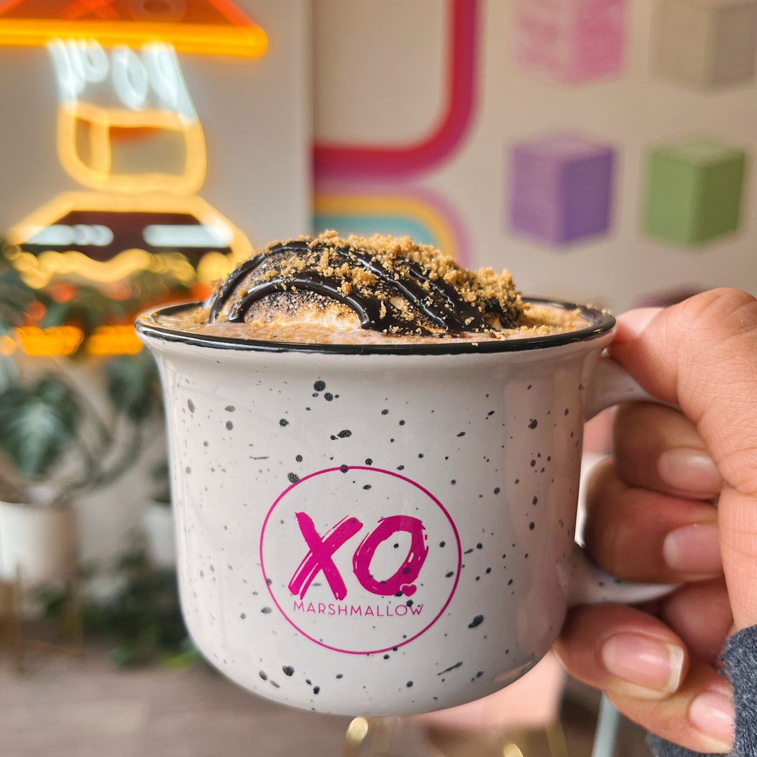 A ceramic campfire-style mug with a white base and black speckles, featuring the 'XO Marshmallow' logo in pink. The mug contains a scoop of Vanilla ooey marshmallow goodness on top of hot chocolate with a drizzle of chocolate ganache and a sprinkle of gluten free graham crackers. The mug is perfect for XO Marshmallow fans.
