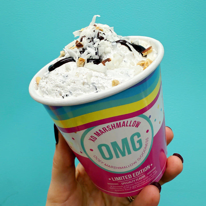 Hand holding a colorful marshmallow fluff container with 'OMG' branding against a blue background