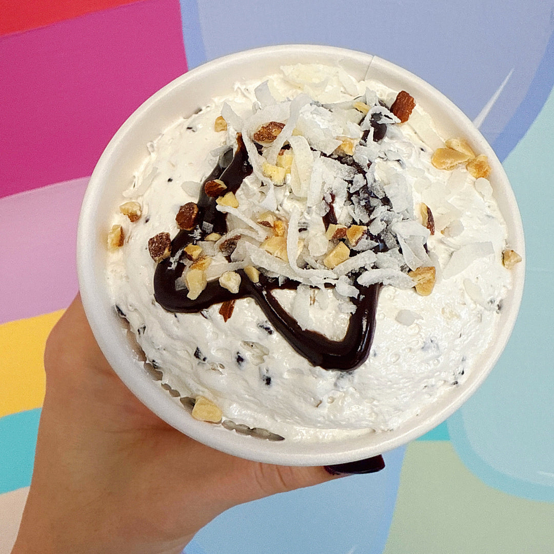 Hand holding a cup of dessert with whipped marshmallow creme, chocolate drizzle, and nuts against a colorful background