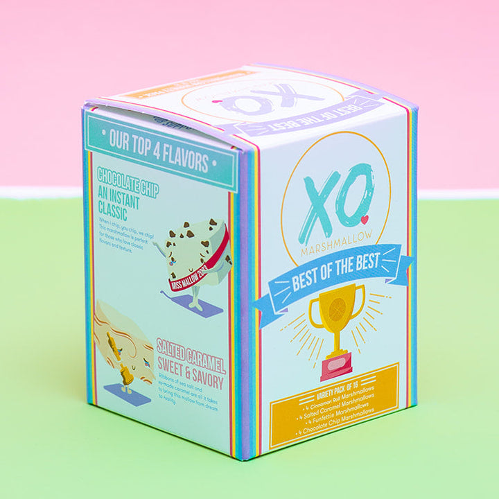 A small branded product box standing upright on a pastel pink and light green surface. The box is mostly white with blue, teal, yellow, and orange printed graphics and text, including a large 'XQ' logo and a trophy illustration.