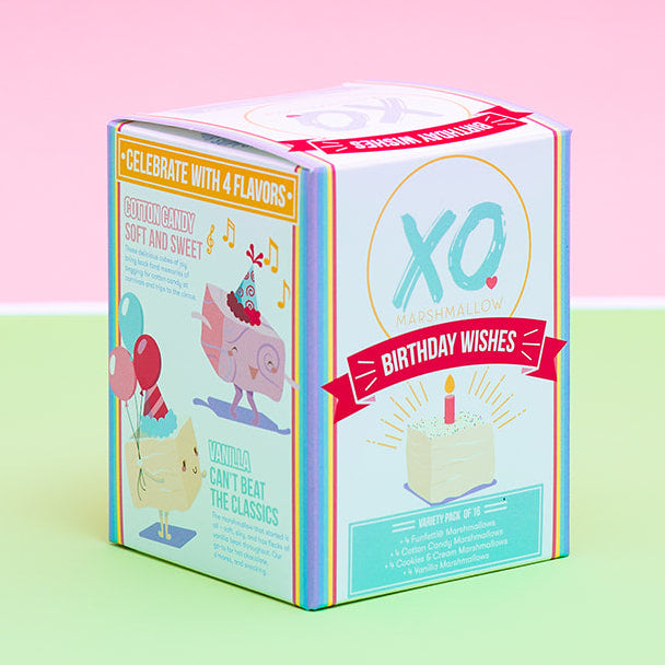 A small illustrated product box standing upright on a pastel green surface against a pink background. The box has colorful graphics, including a large 'XO' logo and birthday-themed illustrations.