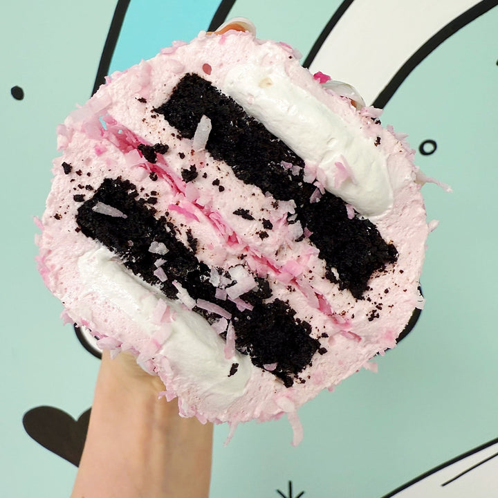 A hand holding a cross section of a XO marshmallow bonbon held against a bright blue background, with layers of coconut marshmallow, marshmallow fluff and gluten free brownie visible.