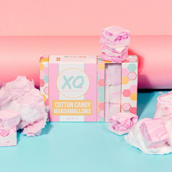 A high end box of XO Marshmallow cotton candy marshmallows against a round pink background with marshmallows and clouds of pink cotton candy around it
