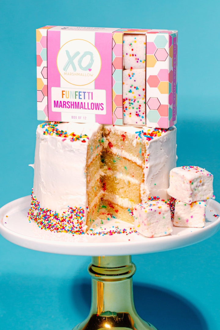 A box of high end Funfetti Marshmallow packaging sitting on top of a birthday cake covered in sprinkles with a large slice missing. The whole thing is on a white and gold cake stand against a blue background