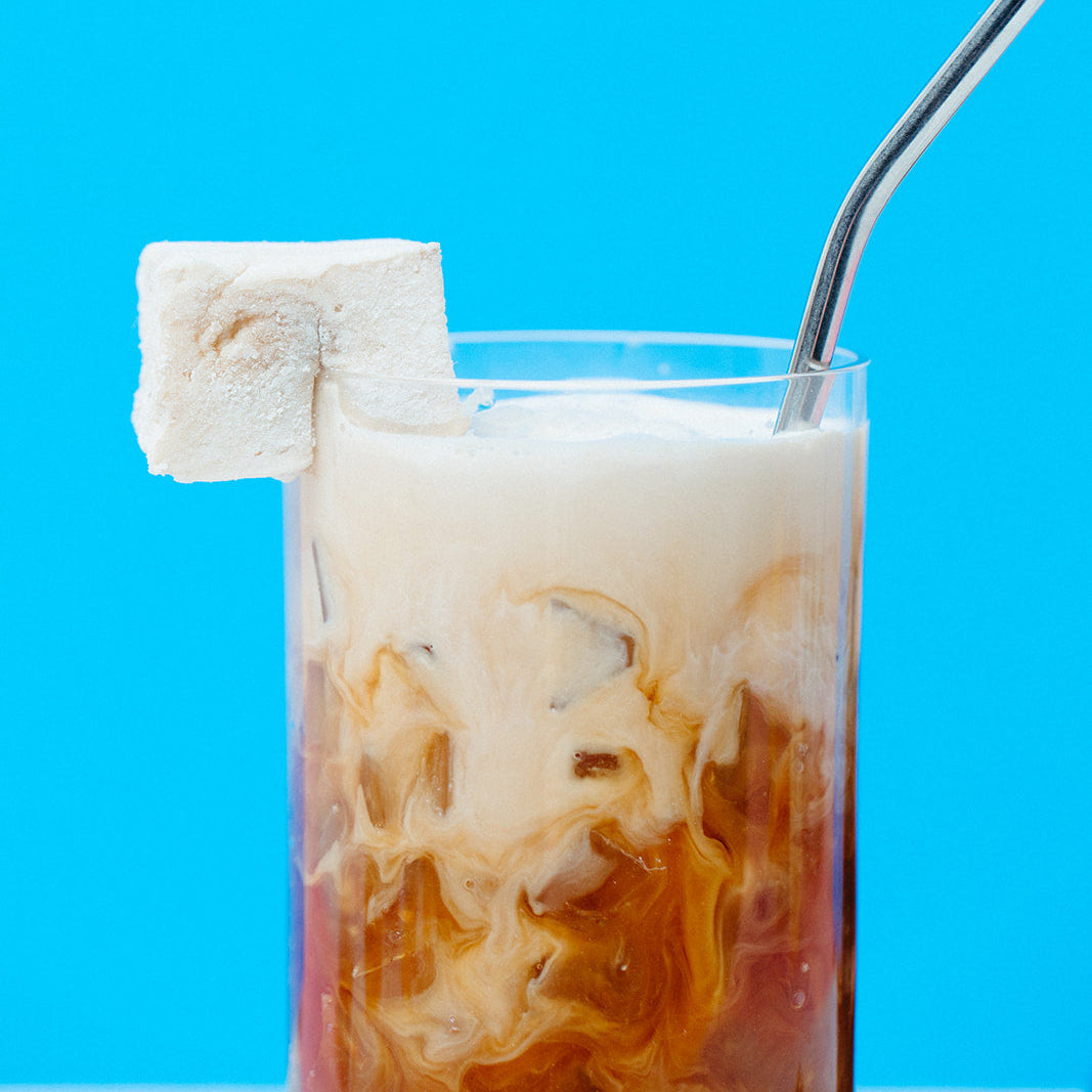 A tall clear glass filled with a layered iced drink, showing dark coffee at the bottom and creamy milk swirls above, topped with a thick white foam layer and a white sugar cube on the rim, with a bent metal straw against a bright blue background.