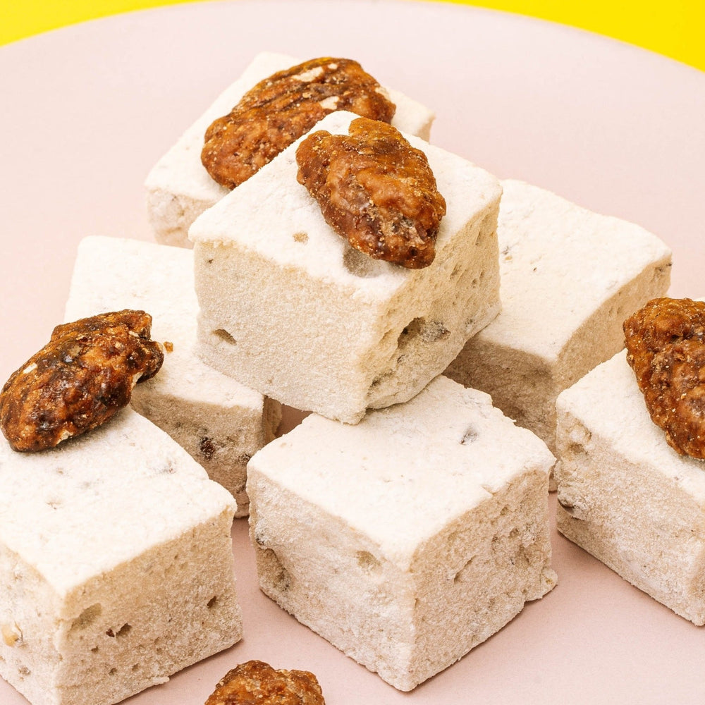 A stack of square shaped XO Marshmallow maple pecan marshmallows on top of a light pink plate. There are whole pecans scattered across the top of the pile.