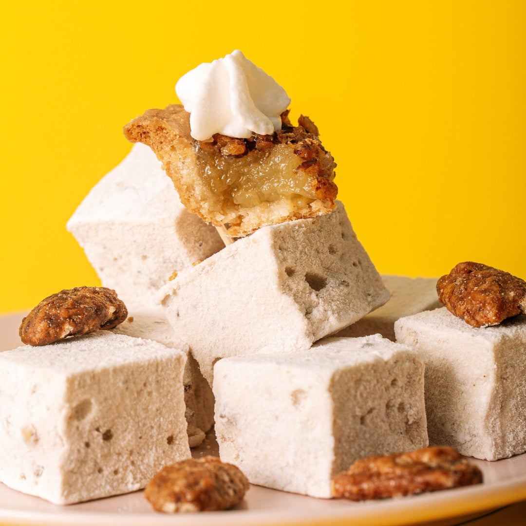 A pile of XO Marshmallow Maple Pecan flavored marshmallows on top of a pink plate with a yellow background. There are whole pecans scattered on top of the marshmallows with a mini slice of pecan pie & whipped cream on top of the pile.