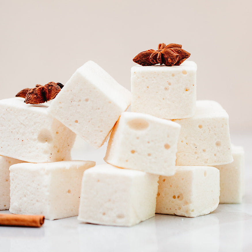 A stack of white marshmallows, topped with brown star anise, arranged on a light marble surface against a soft beige background.