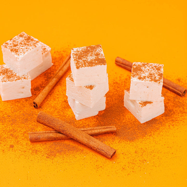 Marshmallow sweets dusted with cinnamon powder, arranged with several cinnamon sticks on a bright orange background.