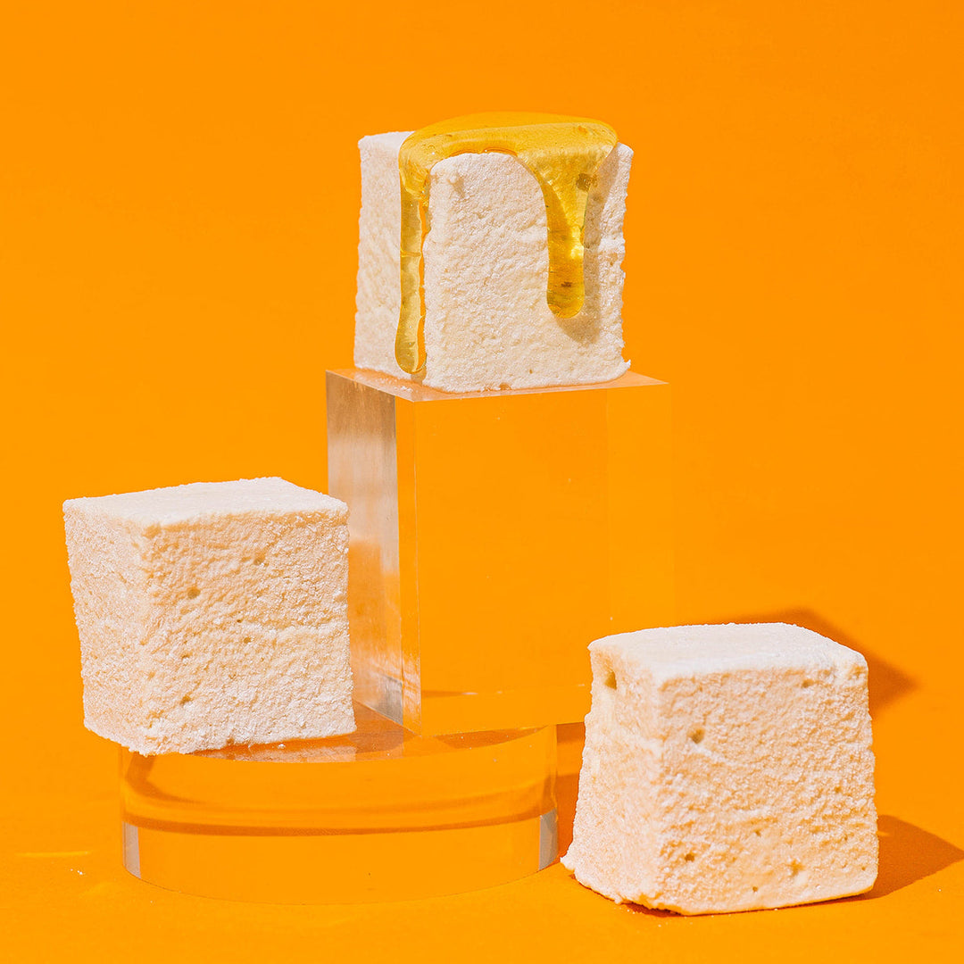 Three white marshmallow cubes displayed on clear acrylic pedestals against a bright orange background. The top marshmallow is topped with a maple drizzle