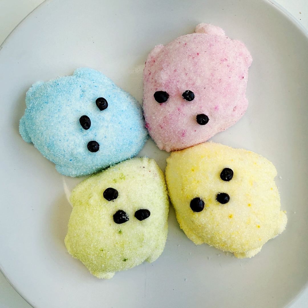 Four large marshmallows shaped like chicks laying on a plate. One is blue, one is pink, one is green, one is yellow. They all have chocolate eyes and beaks.