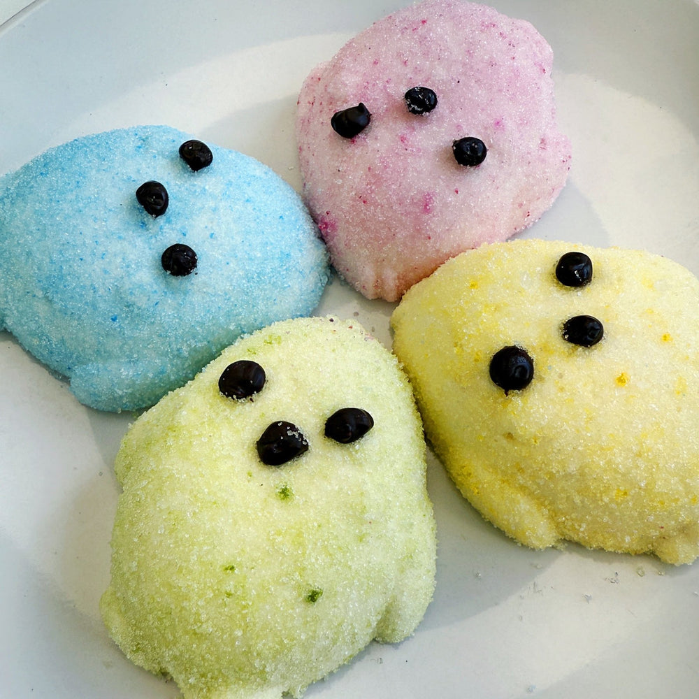  Four large marshmallows shaped like chicks laying on a plate. One is blue, one is pink, one is green, one is yellow. They all have chocolate eyes and beaks.