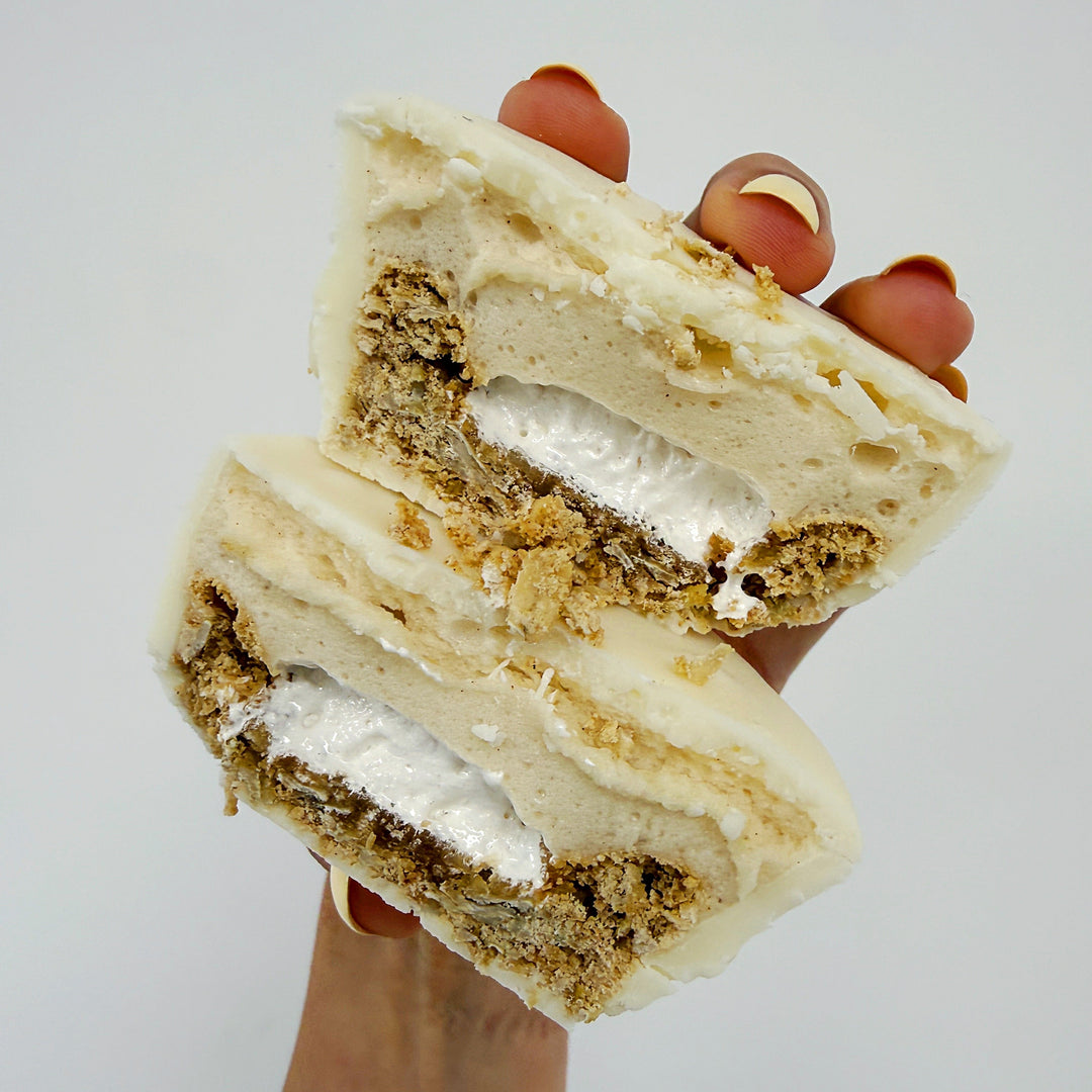 A hand holding two halves of a layered dessert, showing cream-colored frosting, a white cream filling, and a crumbly oatmeal cookie, set against a plain light gray background.