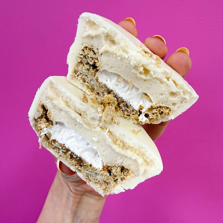 A hand holding a cut open white frosted pastry or bagel with a crumb-topped filling, set against a solid bright pink background.