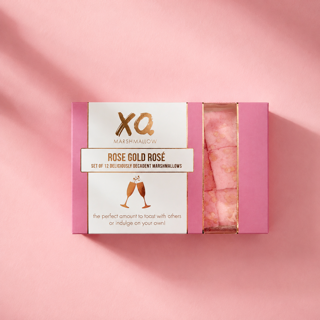 A pink rectangular box of XQ Marshmallow 'Rose Gold' decadent marshmallows, shown open to reveal pink marshmallows inside, set against a matching pink background with soft diagonal shadows.