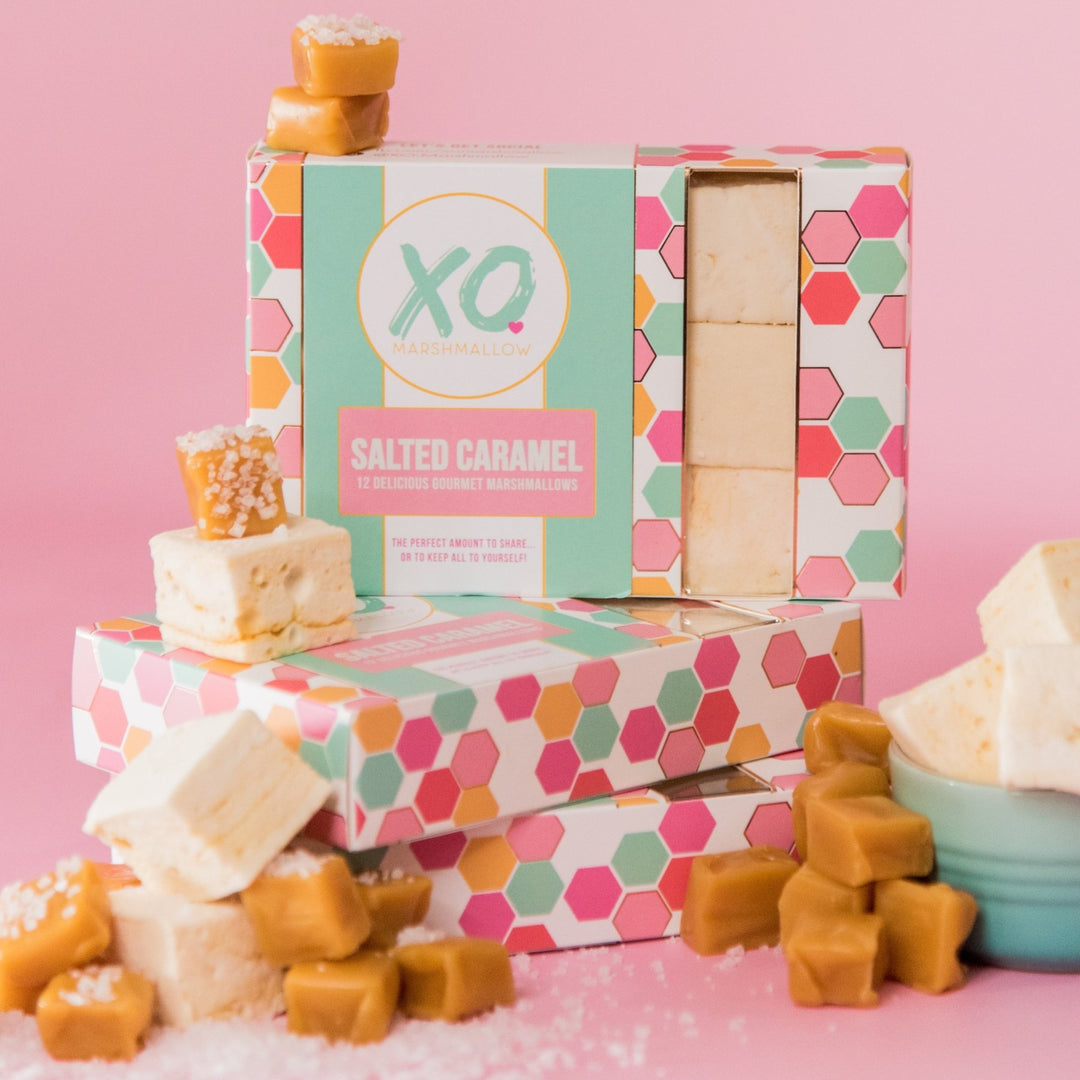 A stack of XO Marshmallow Salted caramel marshmallows inside of their box packaging surrounded by loose salted caramel marshmallows and chunks of salted caramel gooey candy squares.