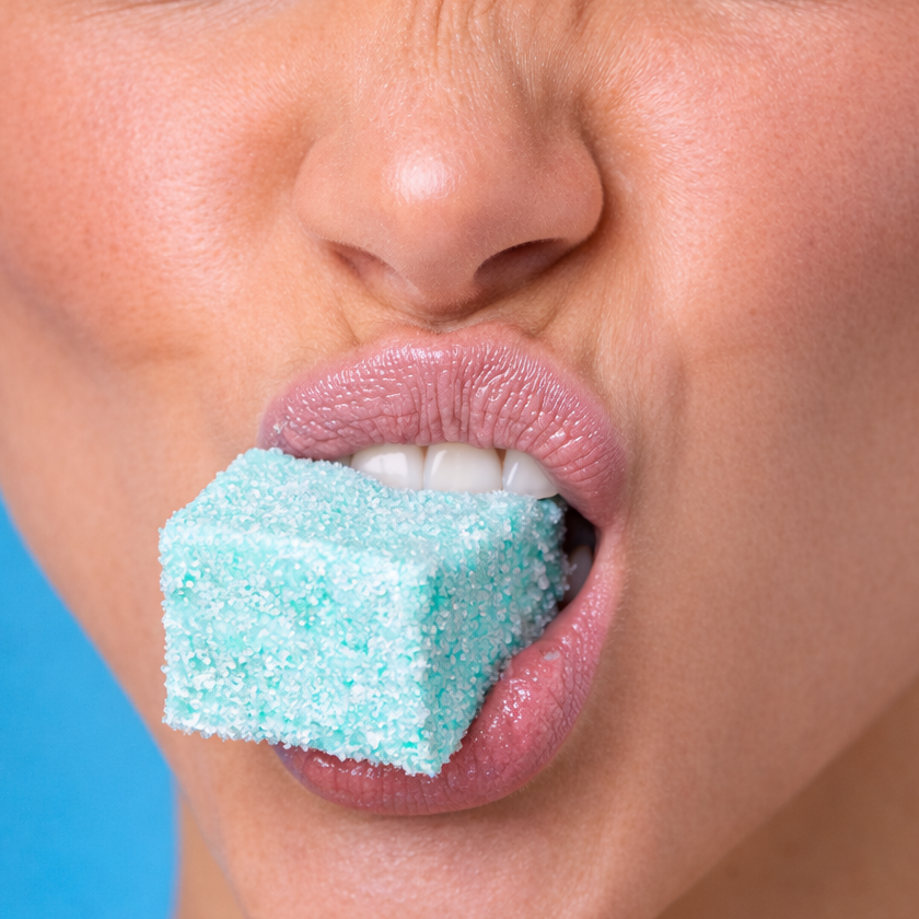 Close-up of a person holding a sour blue raspberry marshmallow in front of their mouth while puckering against a blue background