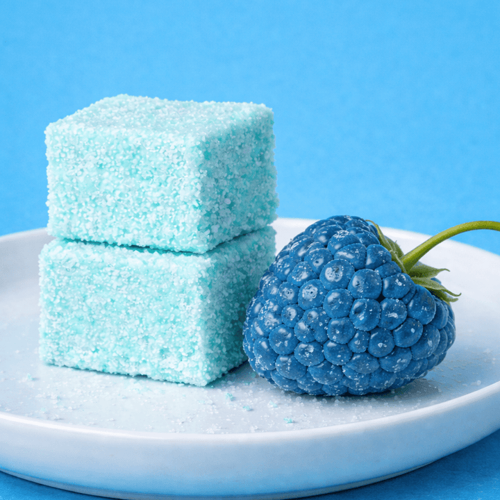 Two sour blue raspberry gourmet marshmallows next to a blue raspberry on a white plate against a blue background