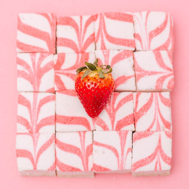 16 XO Marshmallow gourmet strawberry cheesecake marshmallows stacked in a 4x4 grid showing their swirled design with one strawberry sitting on top in the middle of them.