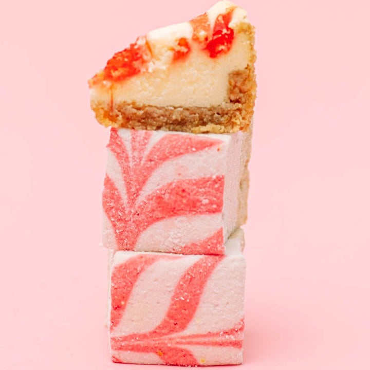 Pink and white swirled xo marshmallows with a slice of cheesecake on a pink background