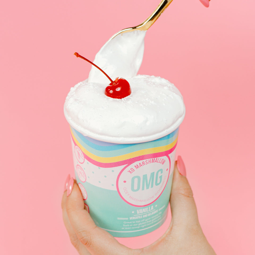 A hand holding a pint of marshmallow fluff with a cherry on top, against a pink background. The cherry is just in the photo as decor and will not be included when it is shipped to you.