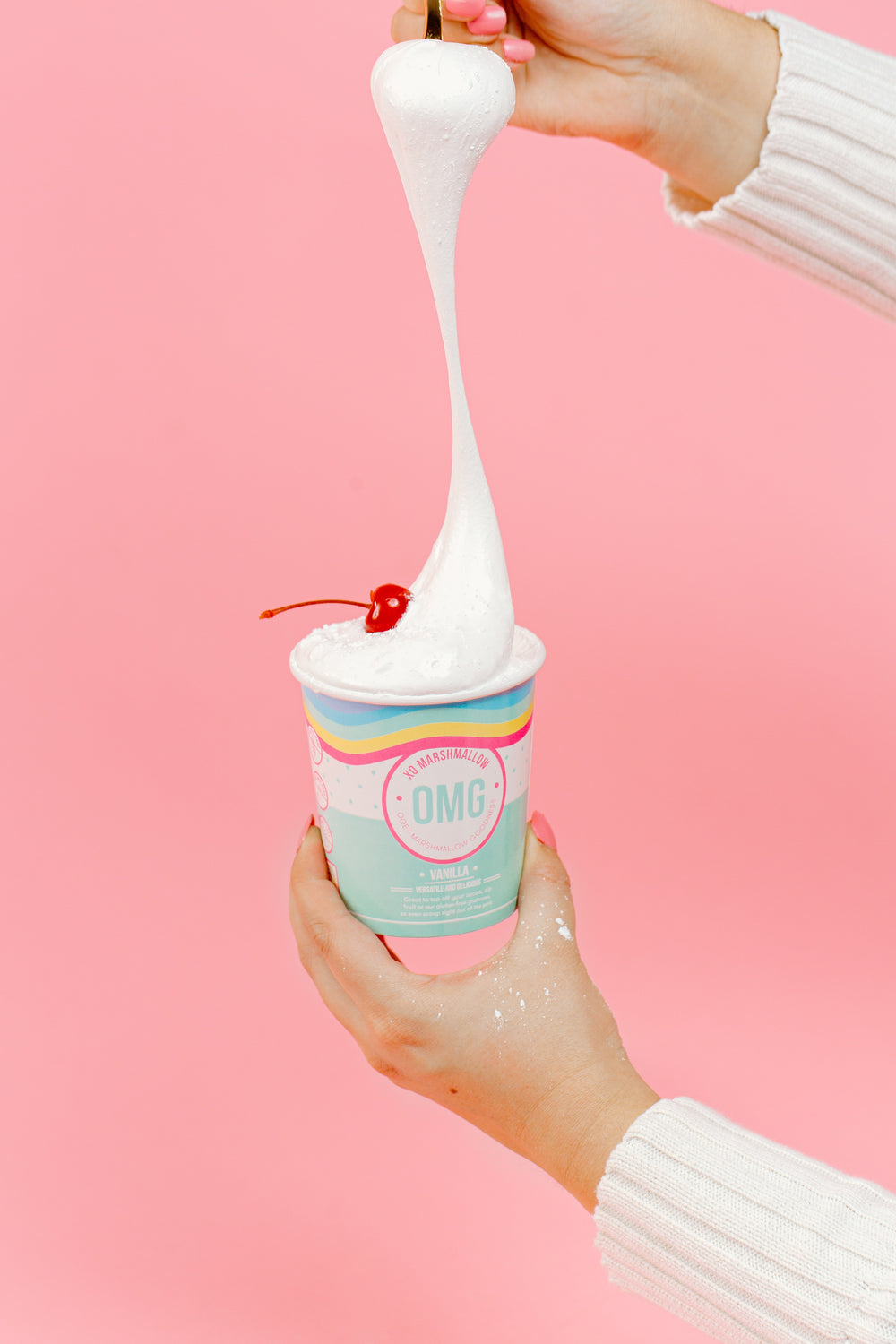 A hand holding a pint of XO Marshmallow vanilla ooey marshmallow goodness in one hand and stretching a spoon full of marshmallow fluff from the pint to the top of the photo with a spoon in the other hand. You can see this person's white sweater sleeves. It is against a pink background.