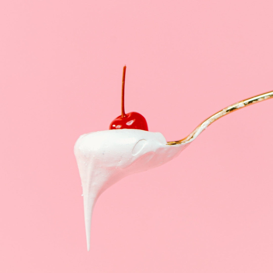 A gold spoon with a scoop of XO Marshmallow Vanilla Marshmallow fluff dripping off of it. There is a cherry on top of it. The cherry is only for the photo and not part of the product. It's all against a light pink background.