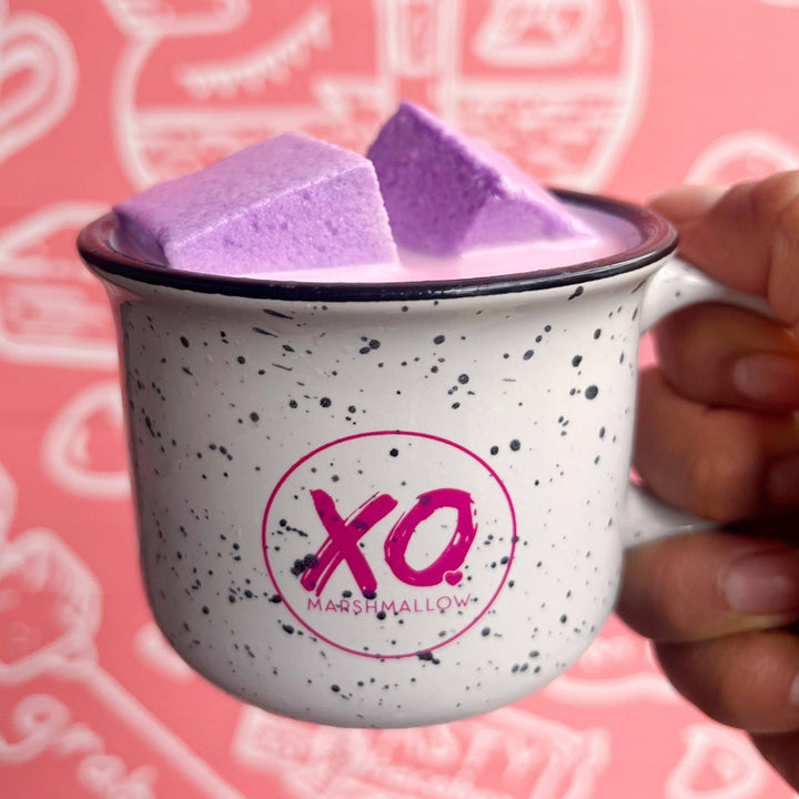 A ceramic campfire-style mug with a white base and black speckles, featuring the 'XO Marshmallow' logo in pink. The mug contains purple marshmallows and is held against a pink background with doodle-like graphics.