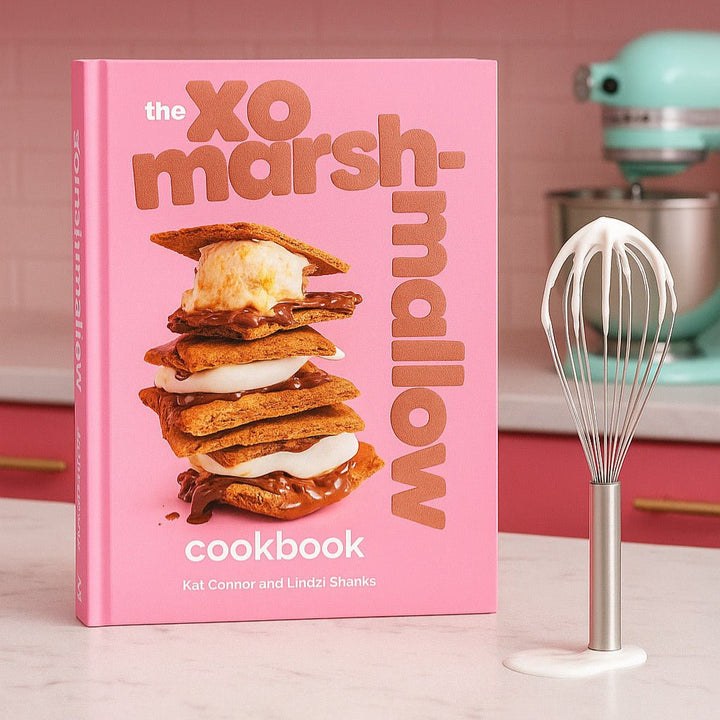 Pink cookbook titled 'XO Marshmallow Cookbook' with a stack of marshmallow treats on a kitchen counter.