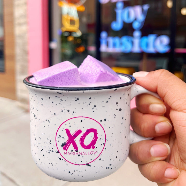 A ceramic campfire-style mug with a white base and black speckles, featuring the 'XO Marshmallow' logo in pink. The mug contains purple marshmallows and is held against the backdrop of the world's first marshmallow cafe located in Chicago.