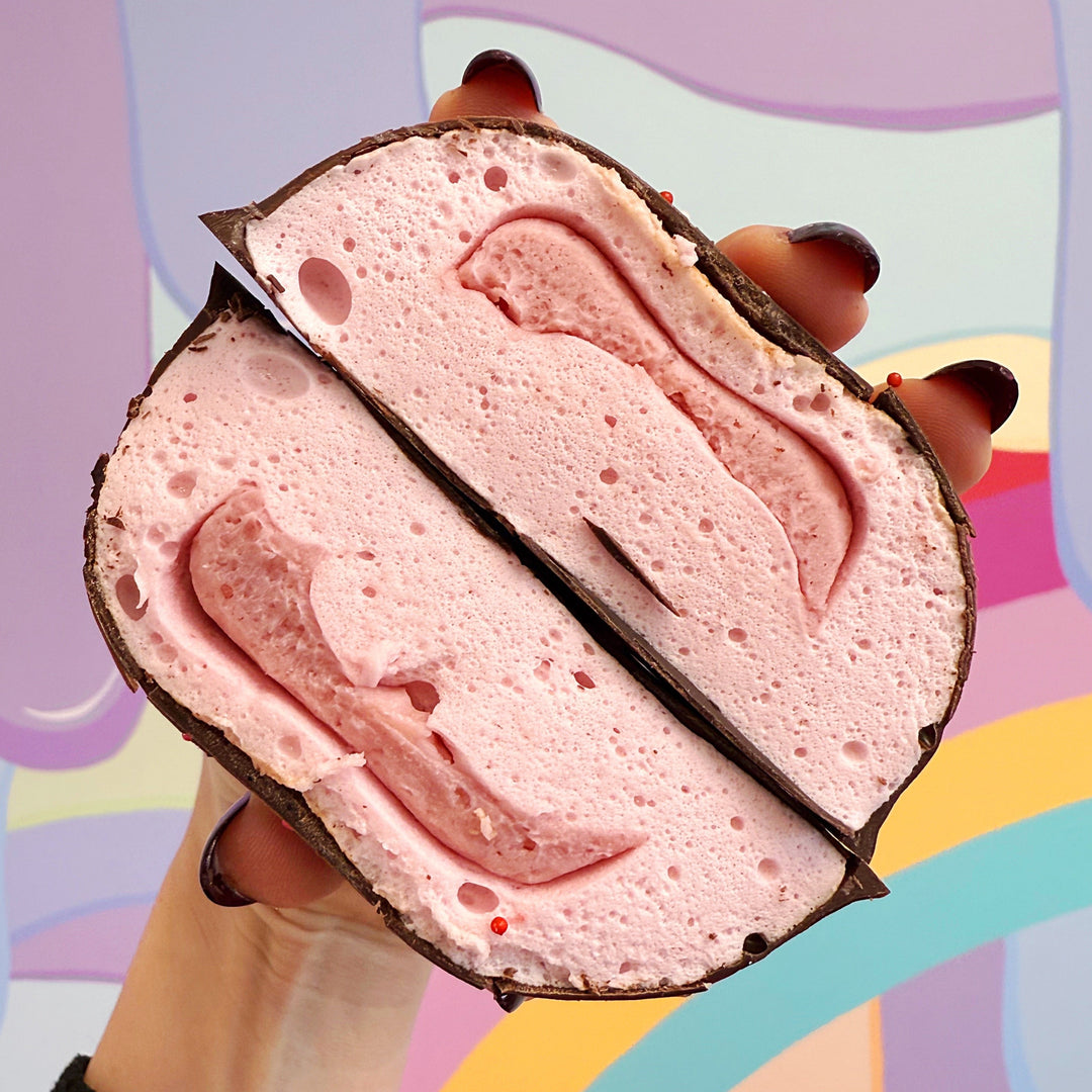 A photo showing a cross section of a strawberry filled XO marshmallow with a chocolate coating and heart-shaped sprinkles on top, held in a hand with a colorful background.