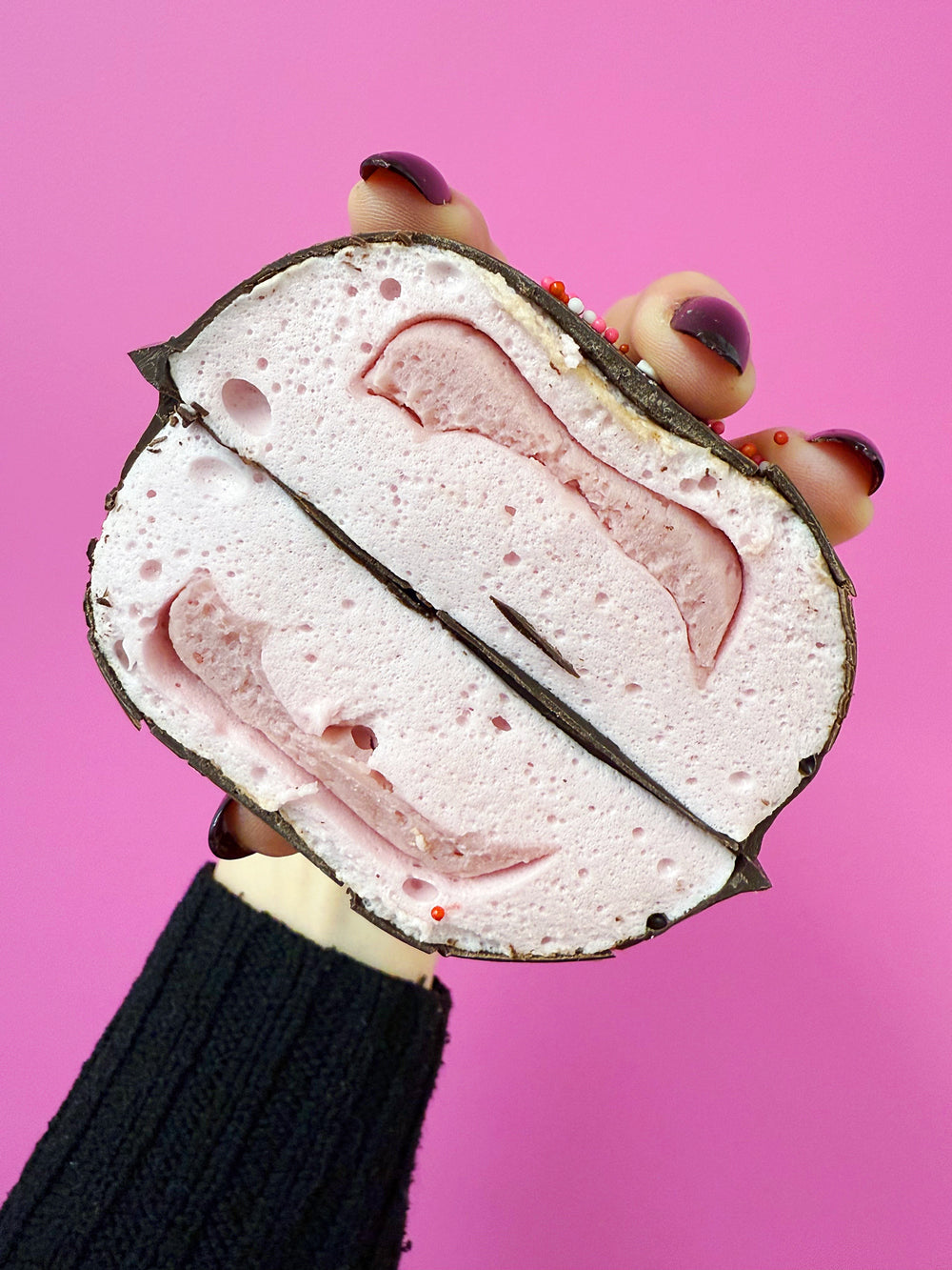 A photo showing a cross section of a strawberry filled XO marshmallow with a chocolate coating and heart-shaped sprinkles on top, held in a hand with a bright pink background. The black sweater of the person holding the treat is showing.