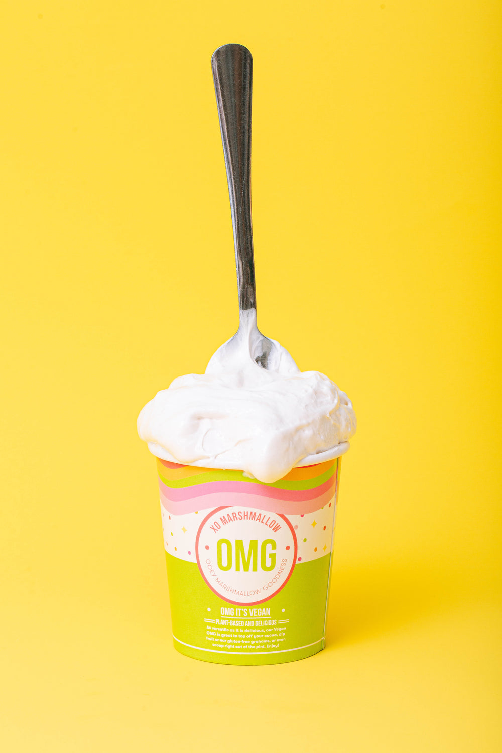 A pint of Vegan Vanilla marshmallow fluff in a lime green ice cream pint with a scoop of vanilla marshmallow creme spilling out over the top with a silver spoon sticking up out of it. All against a bright yellow background.
