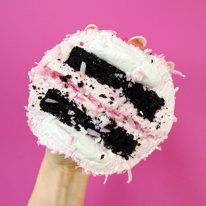 A cross section of a XO marshmallow bonbon held against a pink background, with layers of coconut marshmallow, marshmallow fluff and gluten free brownie visible.