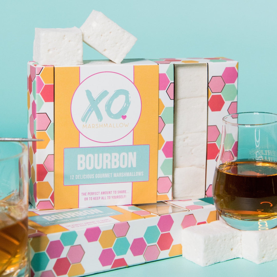 A colorful package of XO Marshmallow with the flavor 'Bourbon' displayed alongside a glass of bourbon whiskey. The marshmallows are presented in a cube shape and the package states each box contains 12 marshmallows.
