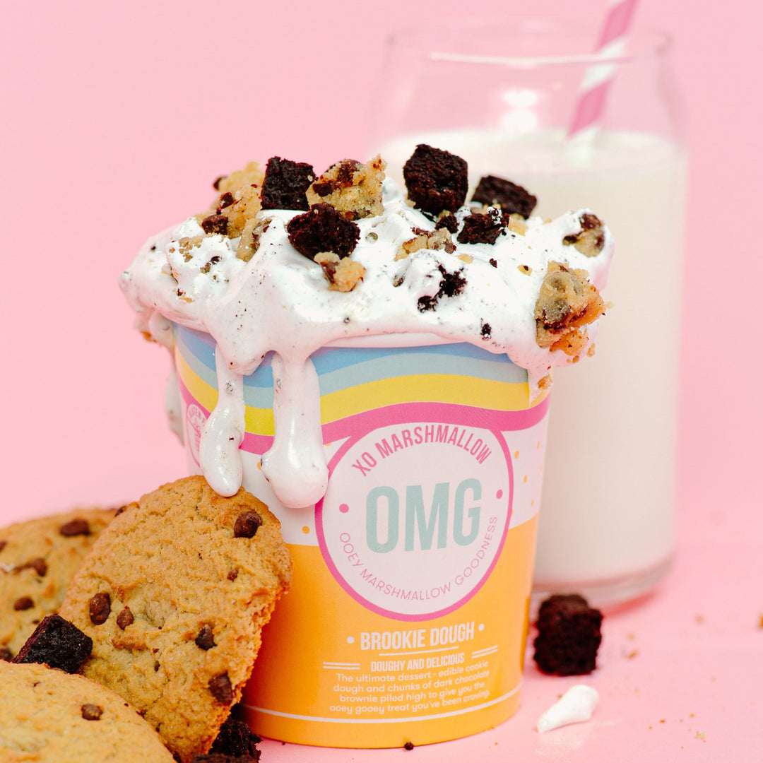 A container of ooey marshmallow goodness with edible cookie dough and brownie chunks next to a glass of milk and cookies on a pink background.