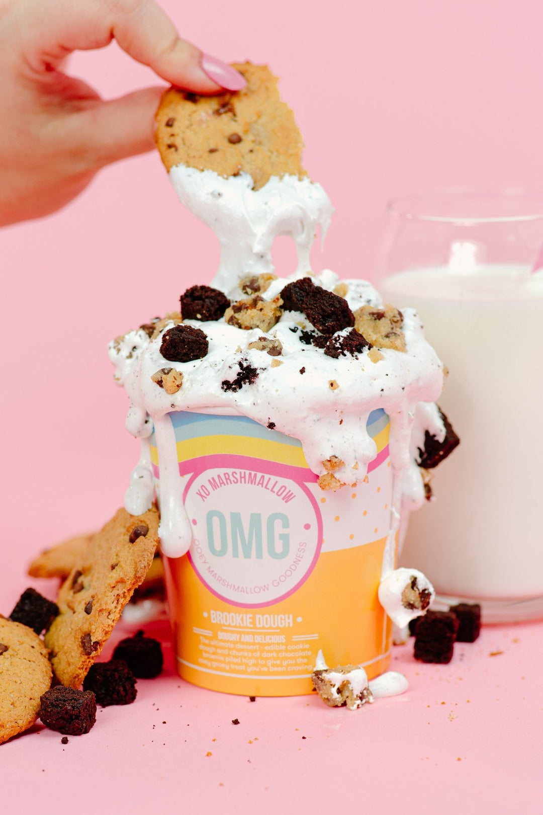 A person dipping a chocolate chip cookie into a pint of XO Marshmallow Brookie Dough ooey marshmallow goodness with chunks of brownie and cookie dough overflowing the pint. There is a glass of milk in the background.