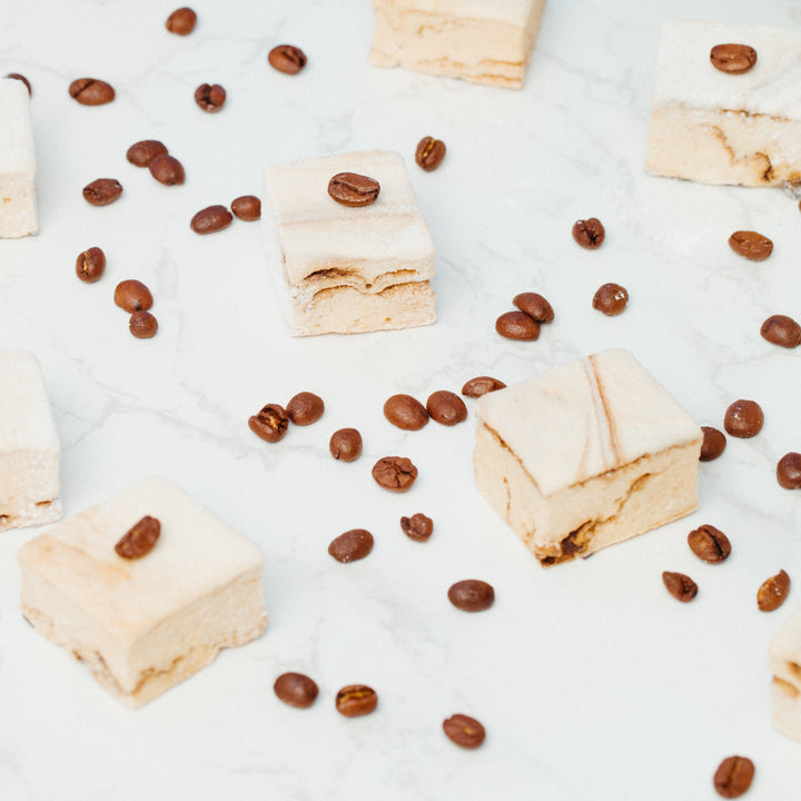 A close up of caffeinated marshmallows against a marble background with coffee beans scattered