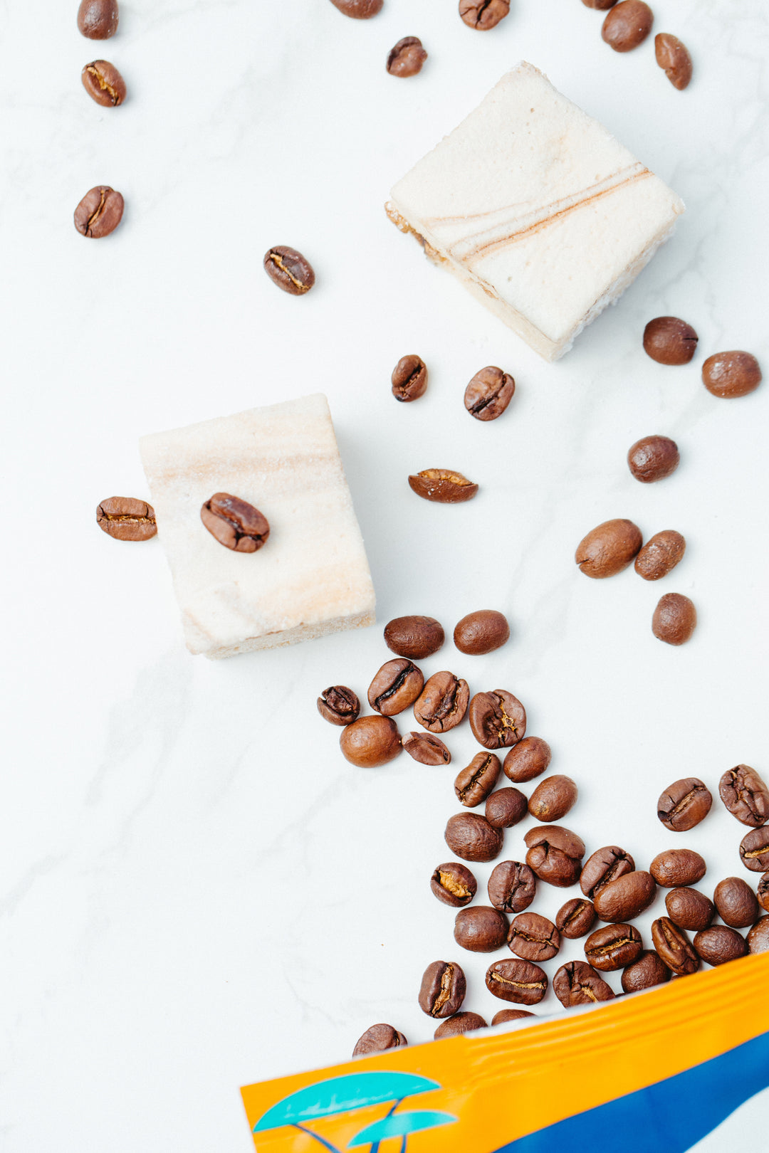 A bag of coffee beans spilled against a marble background with two caffeinated marshmallows among them