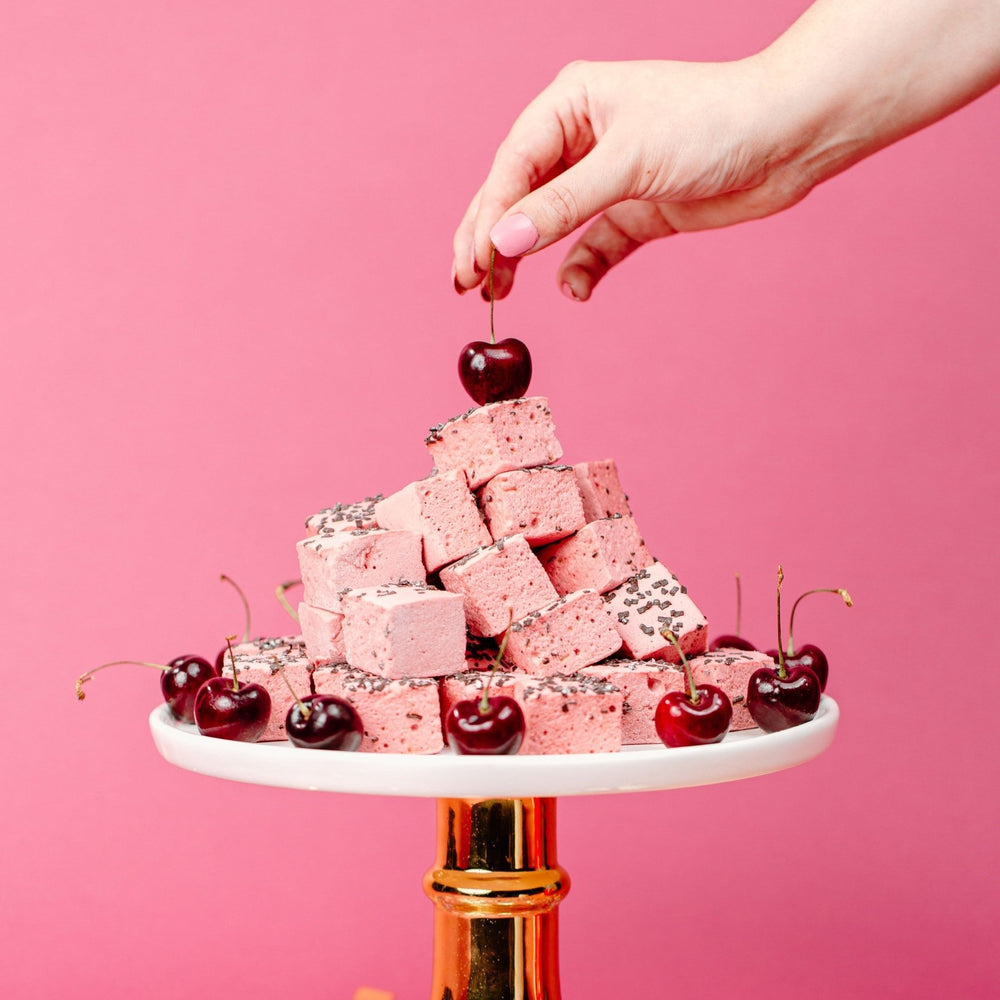 Cherry Cordial Marshmallows piled high on a cake stand while someone holds a tart cherry on top of them
