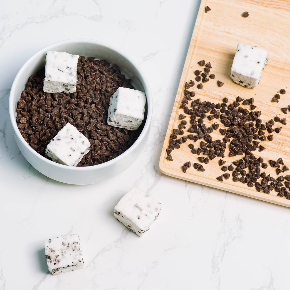 A bowl full of chocolate chips with 3 XO Marshmallow chocolate chip marshmallows in it next to a wood cutting board with chocolate chips scattered on it. All of this is against a white marble background.