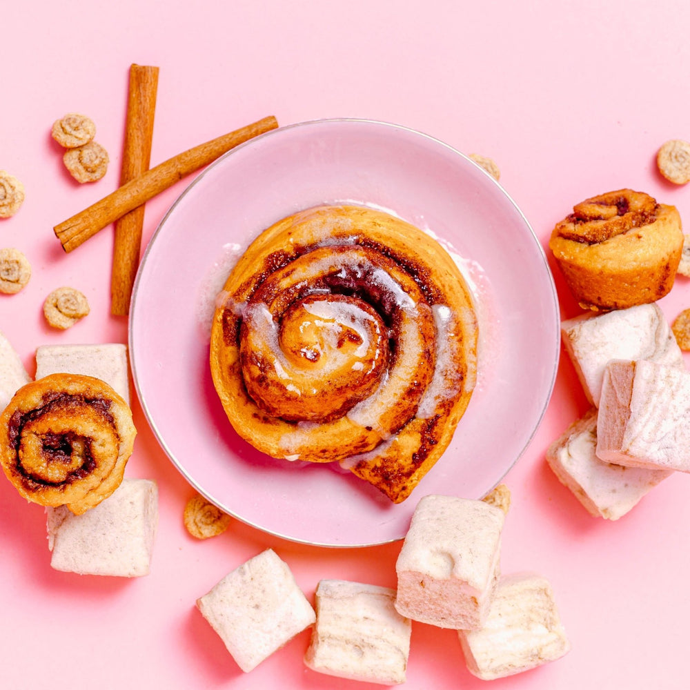 A giant cinnamon roll covered in icing on a pink plate on a pink background. Around the plate are XO Marshmallow gourmet cinnamon roll marshmallows, cinnamon sticks, and mini cinnamon rolls.