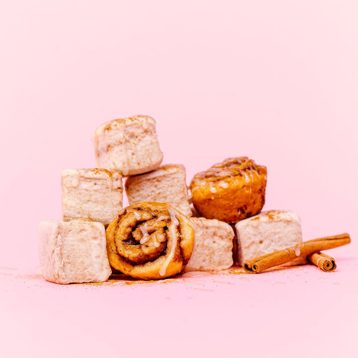 A stack (3,2,1) of cinnamon roll flavored marshmallows with icing drizzled over them. In front of them in a mini cinnamon roll and two cinnamon sticks. All of this is against a light pink background.