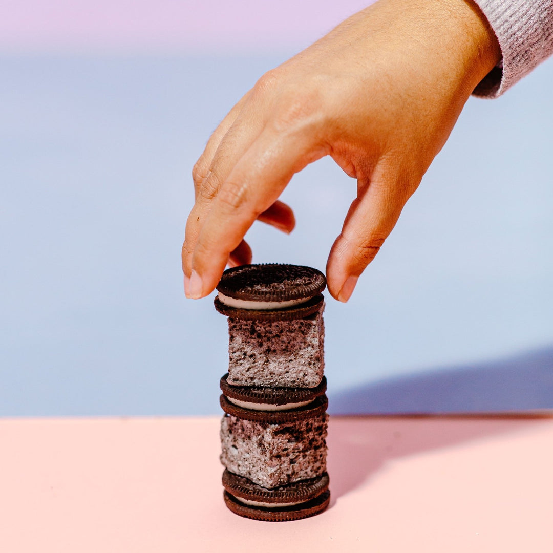 A hand reaching to grab from a stack of alternating XO Marshmallow cookies and cream marshmallows and oreos against a blue background
