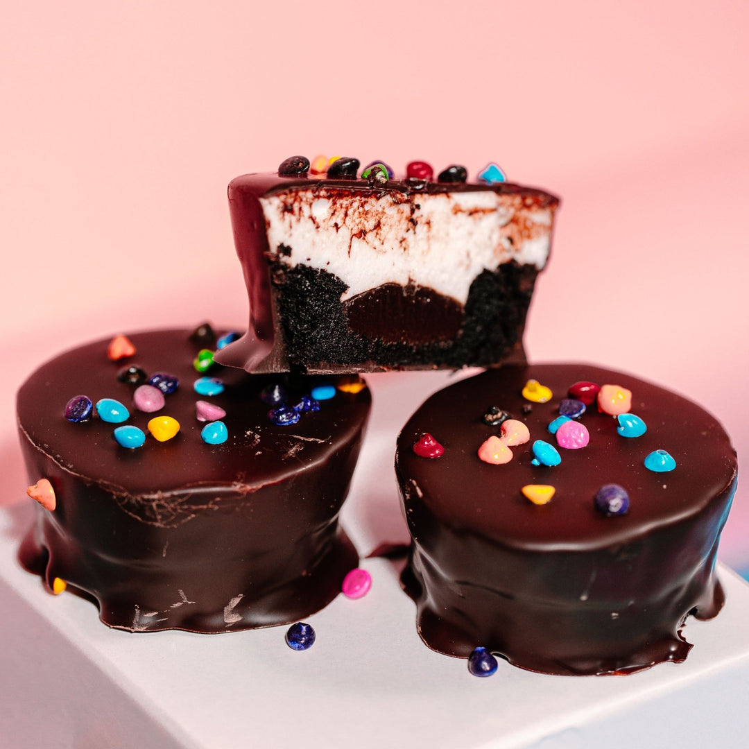 Three XO Marshmallow Gluten Free Gourmet Cosmallow Brownies stacked on top of a white square. Two brownie cups are fully coated in chocolate. The third one on top is a cross section so you can see the brownie, chocolate, and marshmallow inside. All of this is against a pink background. They look like round cosmic brownies.