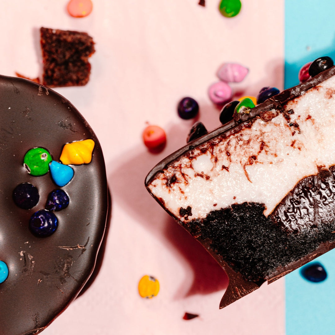 A zoomed in close up of a cosmallow brownie covered in chocolate next to a cross section of one to reveal an inside layer of brownie, chocolate ganache and vanilla marshmallow. 