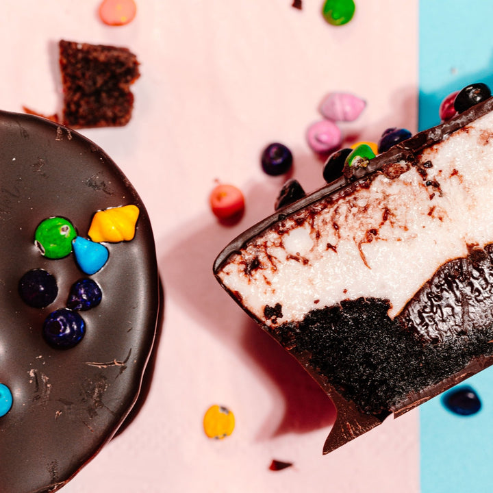 A zoomed in close up of a cosmallow brownie covered in chocolate next to a cross section of one to reveal an inside layer of brownie, chocolate ganache and vanilla marshmallow. 