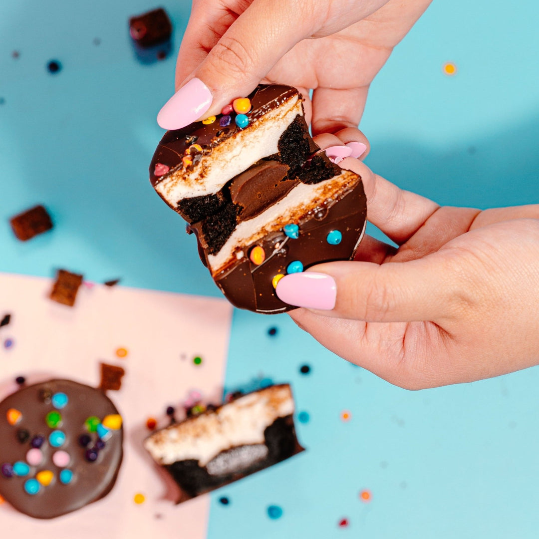 A hand with pink nail polish pulls a XO Marshmallow cosmallow brownie in half so that you can see the inside layers of the treat: brownie, chocolate ganache, and vanilla marshmallow. The outside of it is coated in chocolate and topped with rainbow colorful sprinkles. All of this is against a bright blue background.