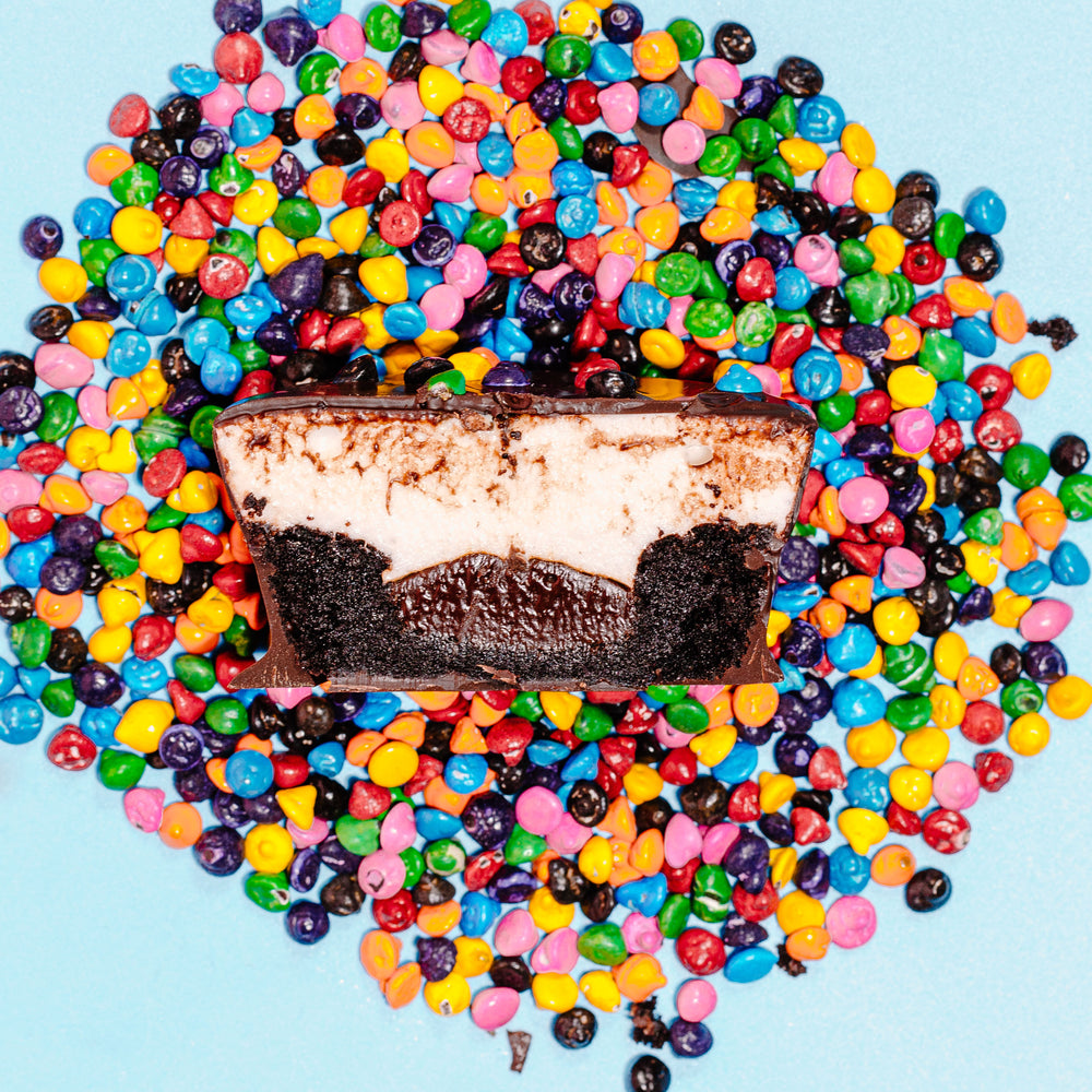 A cross section of an gluten free XO Marshmallow Cosmallow Brownie Cup. You can see the cup is a layer of brownie, chocolate ganache, and vanilla marshmallow with the whole thing dipped in chocolate. All of this is on top of a pile of rainbow chocolate coated sprinkles against a light blue background.