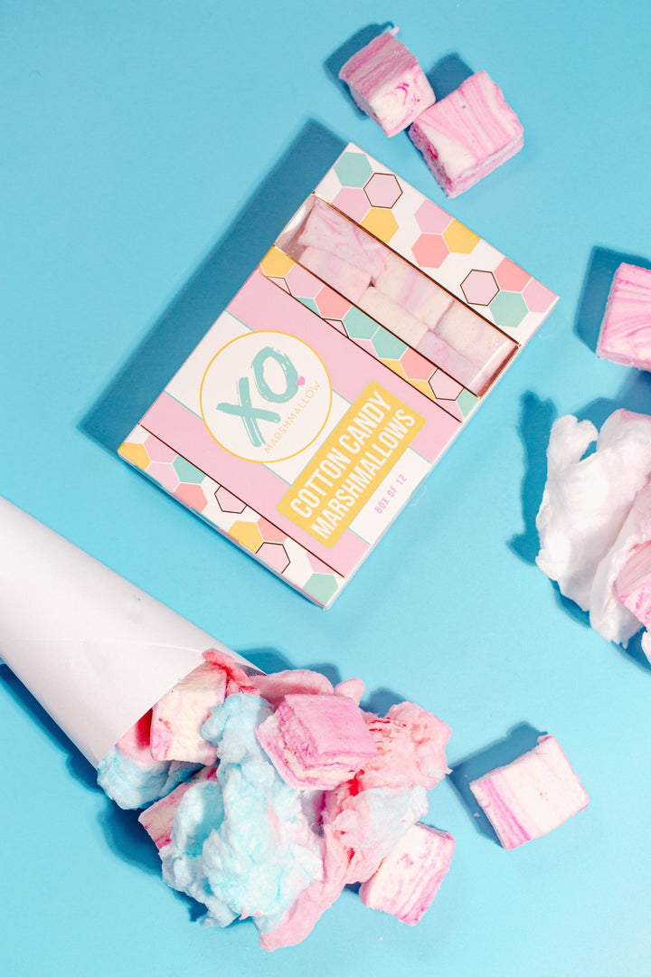 A high end box of XO marshmallow cotton candy marshmallows laying on a bright blue background with cones full of pink cotton candy and homemade cotton candy marshmallows are around it